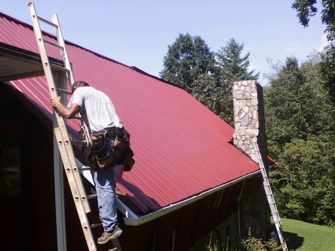 Roofer on a ladder beside a red metal roof and stone chimney — Soffit & Fascia Repair by Roofing Brothers in Baldwin Park